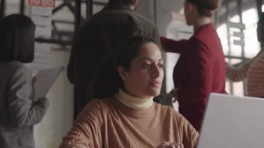 Koyu renk saçlı melez bir kadının göğüs göğsü portatif bilgisayar kullanarak masanın başında oturması, belgelere bakması, arkasında duran tanınmayan meslektaşlarının...