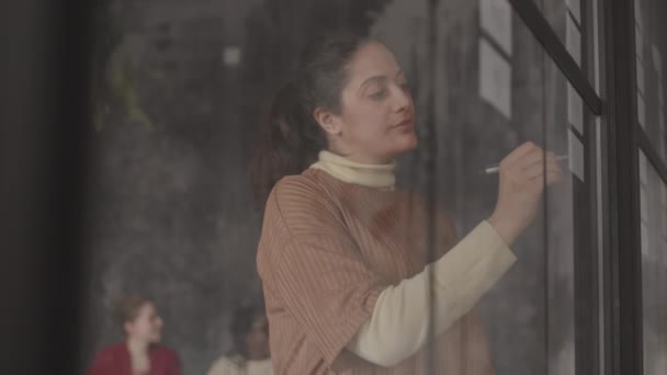 在现代繁忙的办公室里 沉思的混血儿女人手牵着笔 手牵着手 凝视着玻璃墙上的字条 透过玻璃墙向上张望 — 图库视频影像