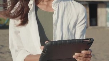 Kulaklık takan, tablet bilgisayar kullanan, bulanık binaların önünde güneşli rüzgarlı sahilde oturan neşeli, beyaz bir kadın.