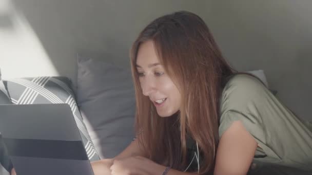 一个快乐的白人女人带着耳机站起来 躺在沙发上 头戴装饰枕头 用笔记本电脑上网 — 图库视频影像