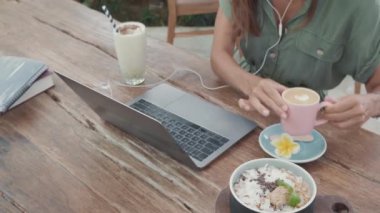 Kafe masasında dizüstü bilgisayarı, bir bardak smoothie, cappuccino ve bir kase yemekle tanınmayan birinin yüksek açılı görüntüsü.
