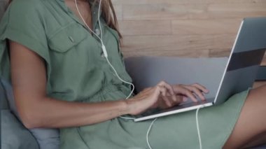 Kablolu kulaklık takan, kapalı bir odada gündüz yatağında oturan, kucağında taşınabilir bilgisayarında video konferansı yapan genç, zayıf beyaz bir kadın.