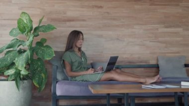 Geniş açılı mutlu beyaz kadın, kablolu kulaklık takıyor, kanepede ayaklarıyla otun içinde oturuyor, kucağında taşınabilir bilgisayarında video sohbeti yapıyor.
