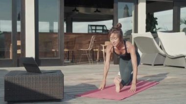 Geniş açıda beyaz kadın spor kıyafetler giyiyor güneşli bir terasta egzersiz paspası üzerinde yoga yapıyor, esneme hareketleri yapıyor.
