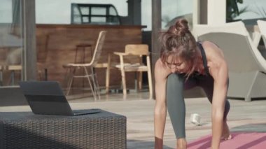 Spor elbiseli beyaz bir kadın, sıcak yaz gününde yoga minderinde esneyerek dizüstü bilgisayardaki mesajlara cevap veriyor.