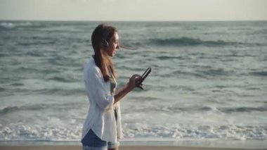 Orta uzunlukta, kahverengi saçlı, kulaklık takan, video çağrısı için tablet bilgisayar kullanan, güneşli, rüzgarlı okyanus kıyısının yanında duran beyaz bir kadın.