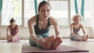 Locked-down of young Caucasian woman wearing sports clothing sitting with straight legs on yoga mat, stretching forward, blurred people exercising on background