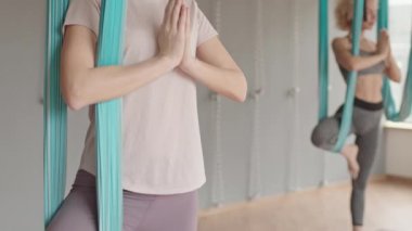 Spor kıyafetleri giyen, bir ayağı kumaş hamakta, elleri namaste içinde meditasyon yapan, başka bir bulanık öğrenciyle meditasyon yapan beyaz bir kadın.