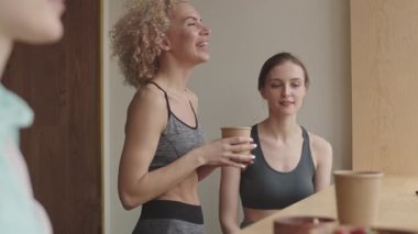 Orta boy, kıvırcık, sarı saçlı, beyaz bir kadın elinde çorba kasesi, el kol hareketleri, kafede arkadaşlarıyla konuşuyor.