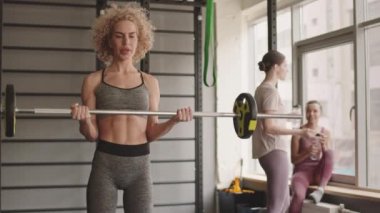 Orta boylu, sarışın, beyaz kadın sporcu spor kıyafeti giymiş halter bisepsi egzersizi yapıyor, bulanık spor salonu müşterileri su içiyor, konuşuyor, arka planda duruyor.