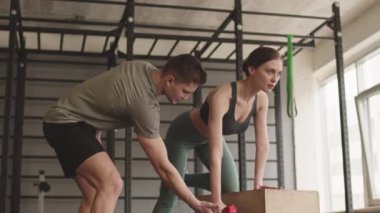Orta uzun mavi gözlü beyaz kadın sporcu spor sutyen ve tayt giyiyor, spor salonunda duruyor, kutuya yaslanıyor, erkek koçun gözetiminde halterle triseps egzersizi yapıyor.