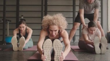 Spor salonunda yoga minderlerinde oturan, öne eğilip, erkek fitness koçunun onlara yardım ettiği üç zayıf beyaz kadının tam pozu.