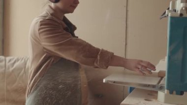 Koruyucu kulaklık ve önlük takmış orta boy genç bir kadın atölyede dikiliyor ve otomatik öğütücüden kalas çıkartılıyor.