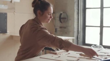 Orta boy kadın marangoz önlük giyer, atölyede tezgah başında dikilir ve oyma tahtalar oyulur.