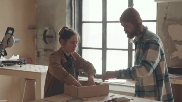 2人の男性と女性の白人の参加者が作業台に立ち 手作りの木製引き出しの仕上げ作業を撮影しました ガラスキャップで箱を覆う男 — ストック動画