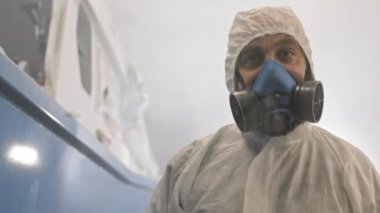 Kapüşonlu ve solunum cihazlı beyaz tehlikeli madde giysisi giyen, dikkatlice bakan, sonra hava fırçası kullanan, kameraya boya sıkan,