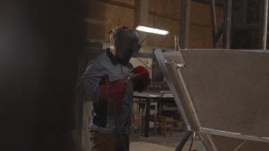 Orta uzunlukta sakallı bir adamın işçi ceketi ve kırmızı eldiven giymesi, kaynak yapan metal bir tekne, sonra kaskını çıkarması ve sonuçlarına bakması.