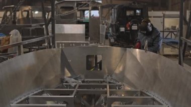 İşçi ceketi ve kırmızı eldiven giyen sakallı bir adam, kaynak yapan metal bir tekne, sonra kaskını çıkarıp tersaneye bakıyor.
