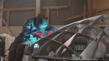 Orta uzunlukta sakallı bir adam, endüstriyel garajda metal iskelet üzerinde oturuyor, kaynak yapıyor, sonra kaskını kaldırıyor ve sonucuna bakıyor.