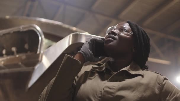 暗い工業用ガレージに立って携帯電話で話している手袋と安全メガネを身に着けている若いアフリカの女性造船所の労働者の低角度胸アップ — ストック動画