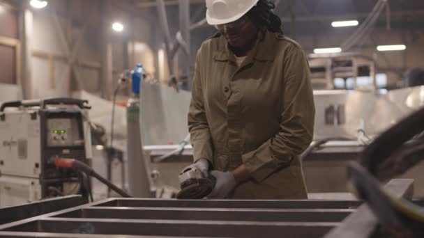産業用ガレージの金属フレームワーク上の研削装置を使用して ハード帽子と手袋を身に着けているアフリカの女性の中程度のショット — ストック動画