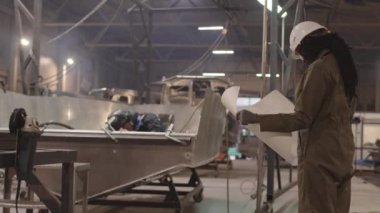 Orta uzunluktaki Afrikalı kadın inşaat işçisi. Sert şapka takıyor ve işçi tulumu giyiyor. Üzerinde taslak olan kağıtlar tutuyor.