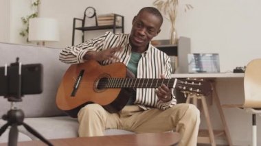 Yetenekli Afrikalı erkek müzisyenin ses tasarım stüdyosunda kanepede oturup akustik gitar çalarken çekilmiş bir fotoğrafı.