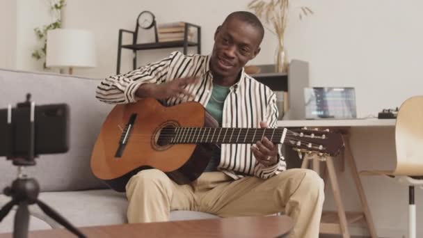 在音响设计演播室里坐在沙发上 在智能手机摄像机上弹奏吉他的有才华的非洲男性音乐家的中景照片 — 图库视频影像