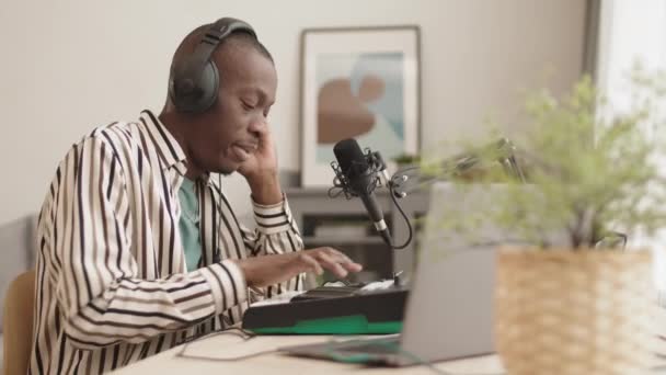中镜头 年轻的非裔美国人头戴耳机 坐在桌旁 一边用麦克风唱歌 一边用笔记本电脑和合成器制作电子音乐 — 图库视频影像