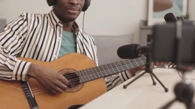 Kulaklık takan, mikrofon ve telefon kamerasının önünde oturan ve akustik gitar çalan Afro-Amerikalı erkek blogcuların kilitlenmesi