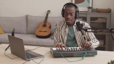 Orta boy Afro-Amerikan erkek sanatçının midi klavye sentezleyicisini çalarken, ev stüdyosunda müzik yaparken dizüstü bilgisayar ve mikrofon kullanarak şarkı söylerken görüntüsü.