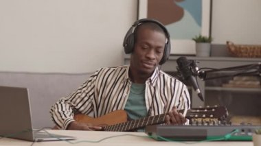Genç Afrikalı-Amerikalı bir erkek sanatçının modern daw 'da oturup mikrofon imzalarken canlı yayın yaparken gitar çalarkenki görüntüsü.