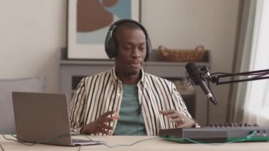 Orta ölçekli genç erkek Afrikalı-Amerikalı müzisyen ev yapımı müzik stüdyosundaki modern daw 'da oturmuş canlı yayın yaparken kameraya bakarken.