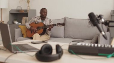 Orta boy Afro-Amerikan müzisyen. Evde müzik stüdyosunda kanepede oturup akustik gitar çalıyor.