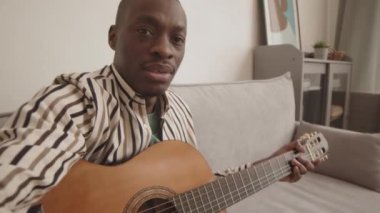 Genç Afro-Amerikan müzisyenin evdeki kanepede oturup akustik gitar çalarken videoda kendini vurduğu orta boy bir fotoğraf.