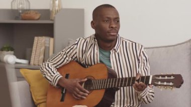 Yakışıklı, kel, Afro-Amerikan müzisyenin evdeki koltukta oturup akustik gitar çalmanın keyfini çıkardığı bir fotoğraf.