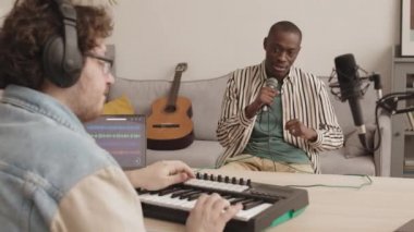 Beyaz adam modern midi klavye sentezleyicisi çalarken, Afro-Amerikalı bir adamın ev yapımı müzik stüdyosunda oturup şarkı söylemesini arkadan izlemek