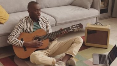 Oturma odasında dizüstü bilgisayarın yanında oturan, taşınabilir stereo hoparlör çalan, akustik gitar çalan ve şarkı söyleyen genç Afrikalı Amerikalı müzisyenleri kilit altına almak.