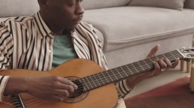 Orta boy, yakışıklı, Afro-Amerikalı bir adamın gündelik giysiler giyip oturma odasında oturmuş akustik gitar çaldığı bir fotoğraf.
