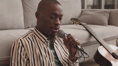 Orta boy yakışıklı Afro-Amerikan erkek müzisyen oturma odasında dizüstü bilgisayarı elinde mikrofonla oturmuş şarkı söylüyor.