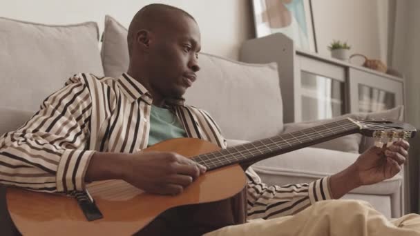 Medium shot of young good-looking African-American man wearing casual clothing sitting on floor in living room and adjusting acoustic guitar