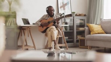 İyi görünümlü Afro-Amerikan erkek müzisyen kulaklık takıyor ev ses tasarım stüdyosunda masasında oturuyor ve akustik gitar üzerine yeni bir single yaratıyor.