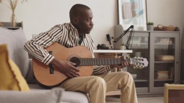 Orta ölçekli Afrikalı genç bir adam gündelik giysiler içinde ses tasarım stüdyosunda koltukta oturup akustik gitar kullanarak melodi yaratıyor.