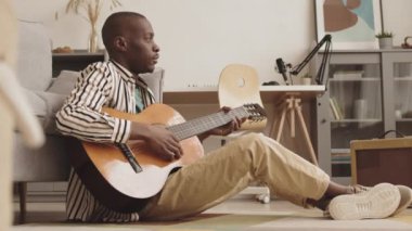 Çekici Afro-Amerikan erkek müzisyenin oturma odasında yere oturup taşınabilir müzik seti hoparlörünün yanındaki kanepeye yaslanarak akustik gitar çalmanın keyfini çıkardığı geniş açı.