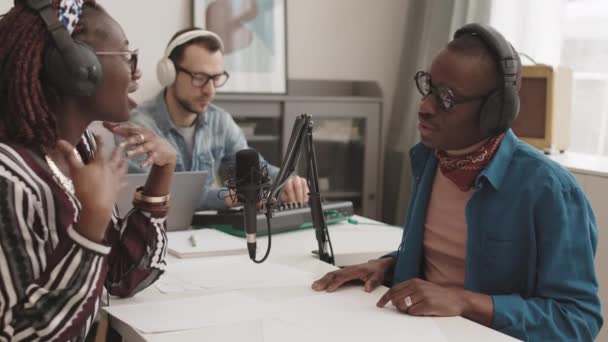 中出しの若いですアフリカの女性ラジオホスト身に着けていますイヤホンでスタジオに座って 彼女の前に座っているスタイリッシュなアフリカ人男性と話をします — ストック動画