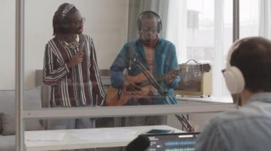Genç, güzel, Afro-Amerikan kadın şarkıcının arka planda şarkı söylerken şık Afrikalı bir adamın profesyonel stüdyoda yeni single kaydı sırasında akustik gitar çalması.