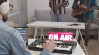 Yetişkin beyaz erkek ses mühendisinin arka planı m-audio keystation 'daki profesyonel stüdyoda otururken müzik grubu yeni albüm kaydederken çalışıyor.