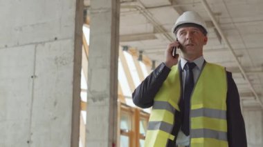 Düşük açılı orta yaşlı beyaz adam yeşil yansıtıcı yelek ve beyaz şapka giyiyor, inşaat alanında cep telefonuyla konuşuyor.