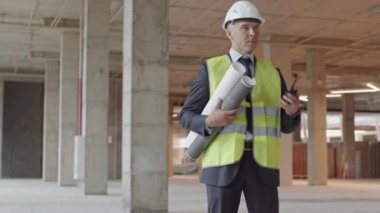Orta yaşlı beyaz bir adamın yeşil yansıtıcı yelek ve beyaz başlık giyip, sarılı planlar taşıyarak, inşaatta telsiz kullanarak orta-uzun yol izlemesi.