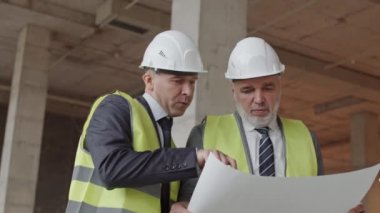 İki orta yaşlı beyaz erkek inşaat işçisi yeşil yansıtıcı yelek ve beyaz başlık giyiyorlar, binada duruyorlar, belgeleri tartışıyorlar.
